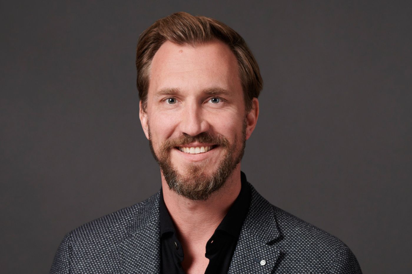 Mann mit kurzem braunem Haar und Bart im dunklen gemusterten Sakko vor neutralem Hintergrund lächelnd porträtiert.