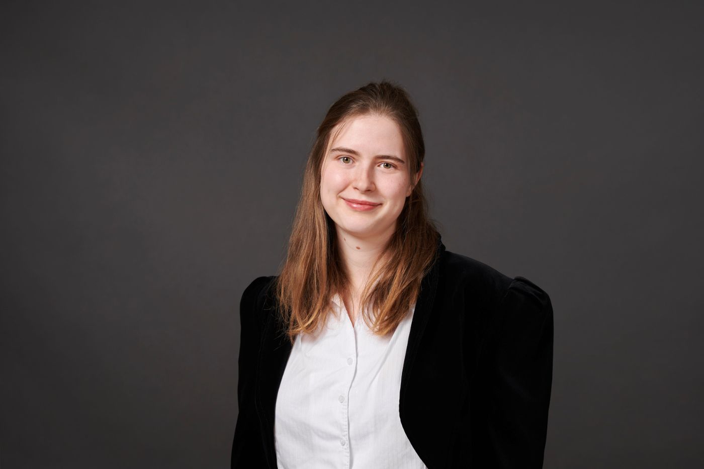 Frau mit mittellangen braunen Haaren in dunklem Blazer und weißer Bluse vor neutralem Hintergrund lächelnd porträtiert.