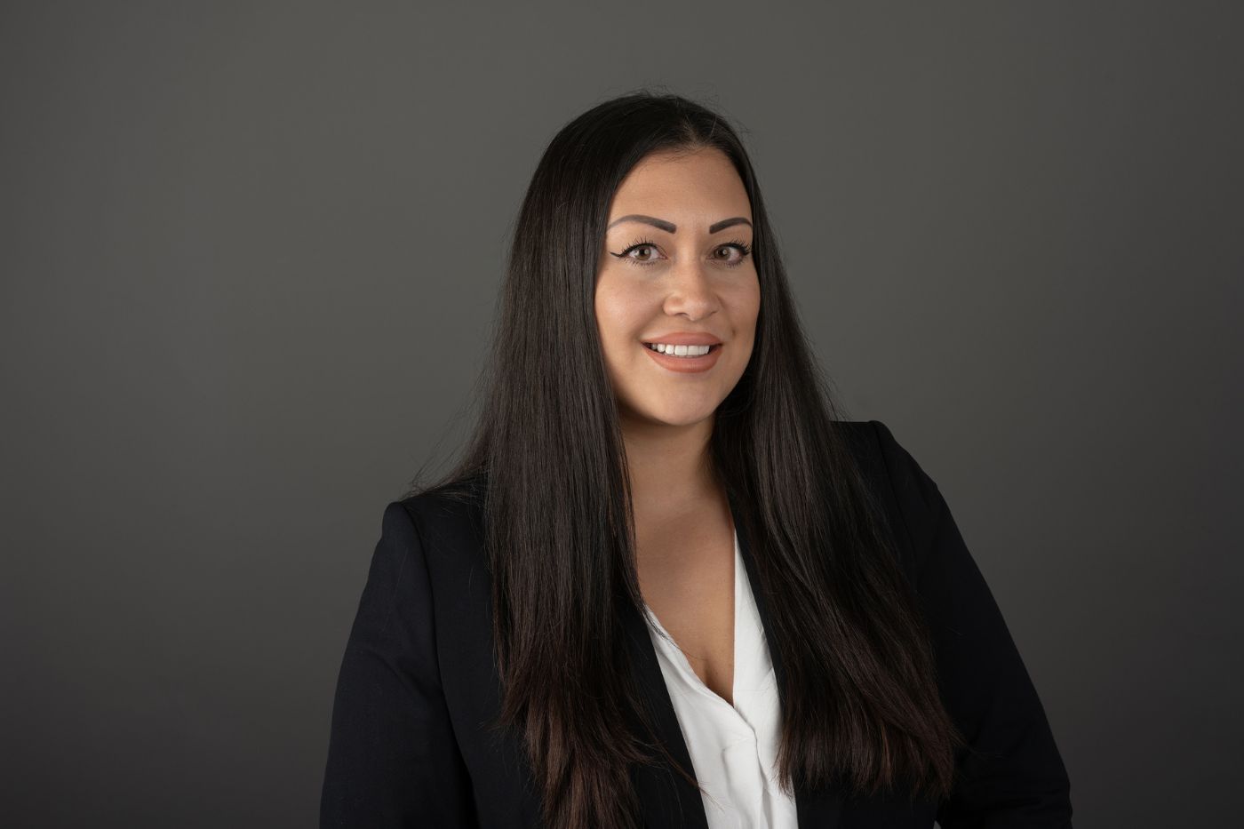 Frau mit langen dunklen Haaren im dunklen Blazer und weißer Bluse vor neutralem Hintergrund lächelnd porträtiert.