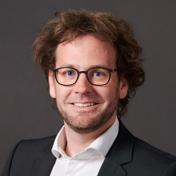 Mann mit Brille und lockigem Haar lächelt im Porträt vor dunklem Hintergrund.